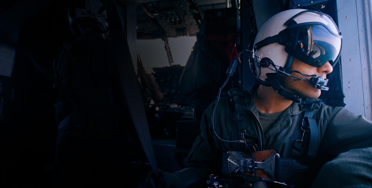 A male warfighter wearing a helmet, eye shield and headset looking out the side of a helicopter